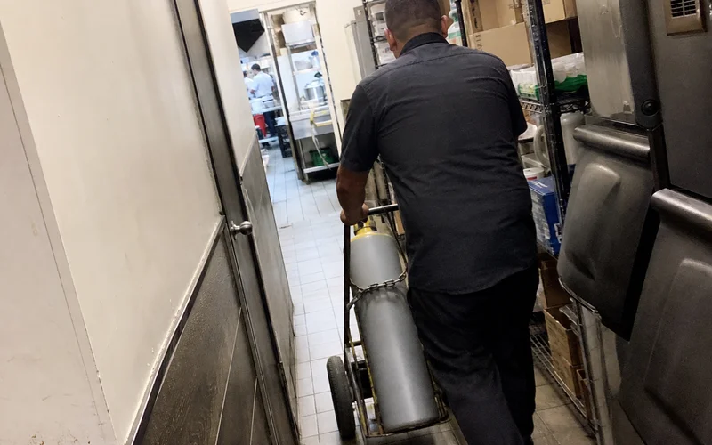 Staff member wheeling a high-pressure gas cylinder through a narrow back hallway