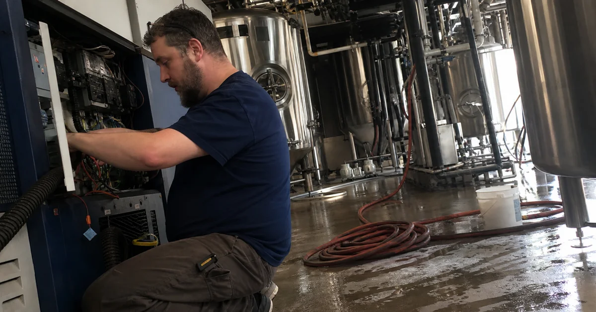 Technician servicing a glycol chiller beside fermentation tanks in a Portland brewery cellar
